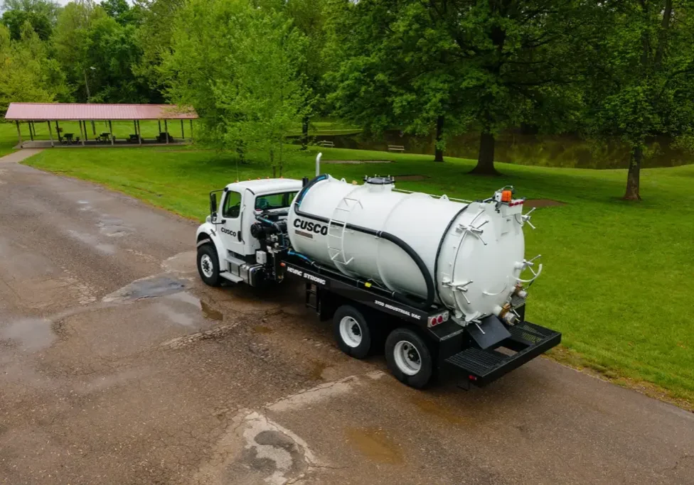 White Cusco vacuum truck with cylindrical tank parked on wet pavement by park pavilion and trees, industrial vacuum equipment